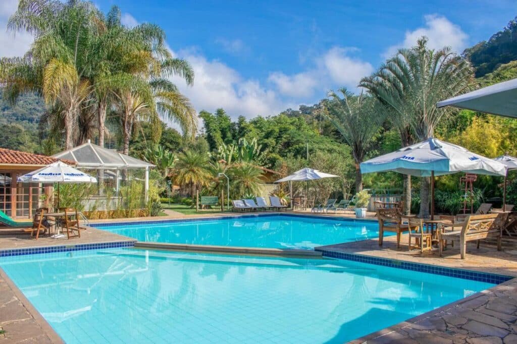 Piscina cercada por palmeiras, espreguiçadeiras e guarda-sóis no Bomtempo Itaipava, um dos hotéis fazenda no Rio de Janeiro. Céu azul claro e um cenário montanhoso.
