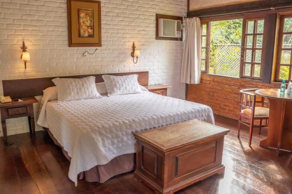 Um quarto aconchegante do Bomtempo Itaipava com uma cama grande, móveis de madeira, paredes de tijolos brancos e uma janela com vista para a vegetação.