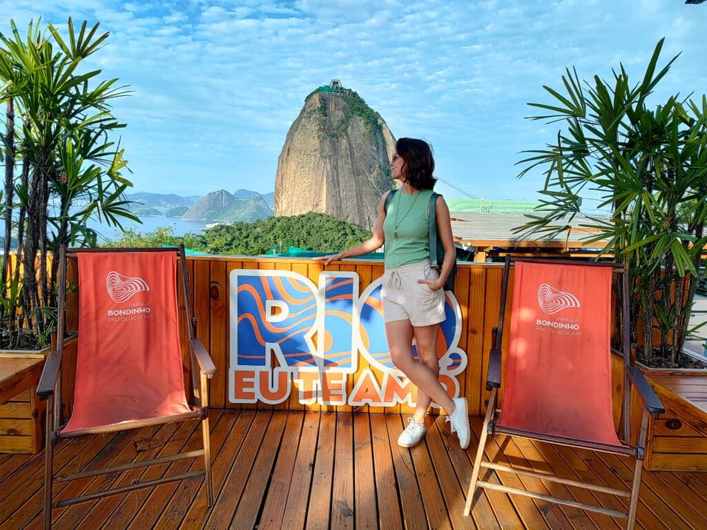 mulher posando para a câmera em frente ao corcovado no Rio de Janeiro. Há duas cadeiras de praia vermelhas de cada lado da moça, ela está olhando para a esquerda, permitindo aparecer somente o perfil do rosto. Imagem para ilustrar post sobre o que fazer no feriado de Corpus Christi.