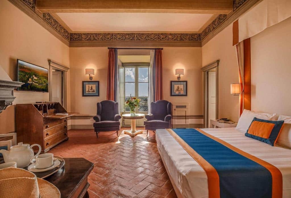 Suíte executiva do Borgo Scopeto Wine & Country Relais, um dos hotéis na Toscana, com 55 m², cama grande de casal, duas poltronas roxas, mesa redonda no meio com flor, janela refletindo a luz do sol e mostrando o céu azul com algumas nuvens, além de móveis de época pelo quarto que tem chão de taco antigo marrom