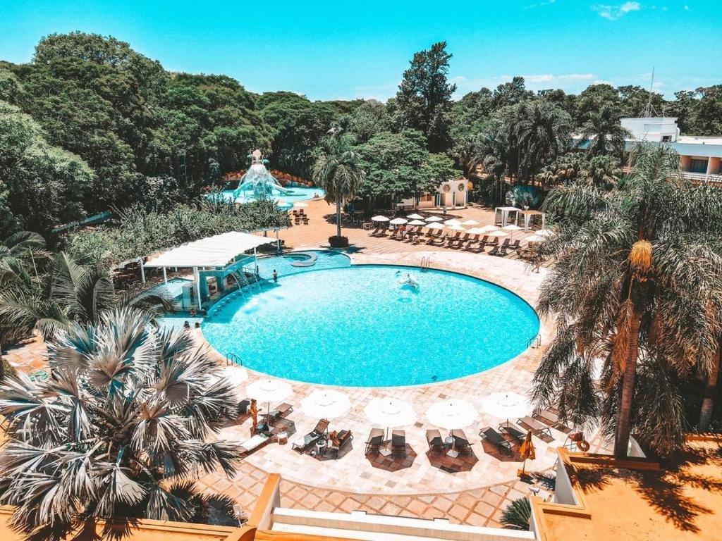 Parte do Bourbon Cataratas do Iguaçu Resort que mostra uma piscina ao ar livre com várias espreguiçadeiras e palmeiras em volta. E ao fundo tem outra piscina e volta das árvores durante o dia. Imagem ilustrando post Resorts em Foz do Iguaçu.