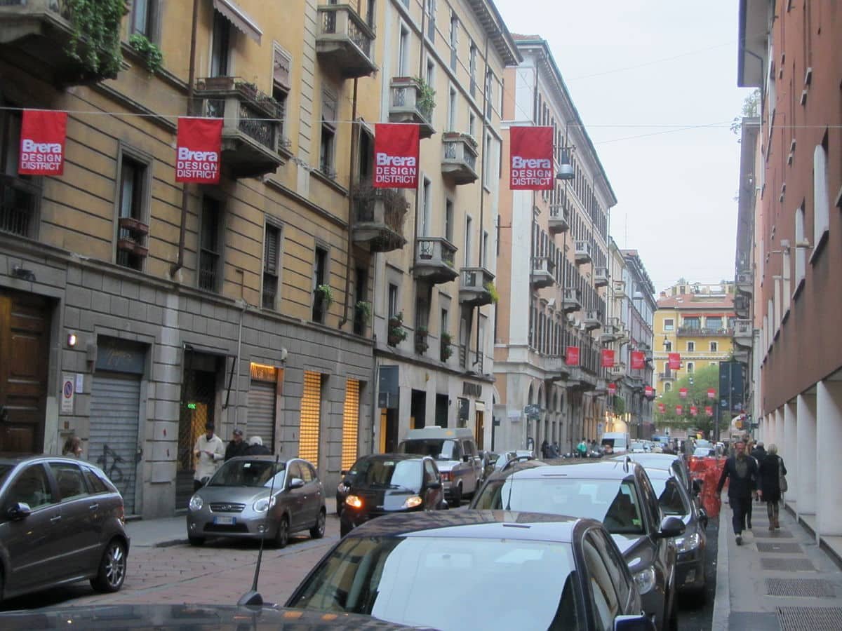 Rua em Brera com carros estacionados ao longo da estrada e placas para Brera Design District. Pessoas andam por prédios históricos.