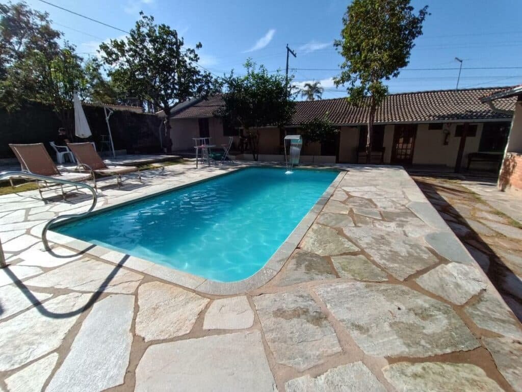 Área da piscina do hotel pousada Brisa da Serra em Pirenópolis. A hospedagem ao fundo, a piscina em primeiro palco, com cascata, cadeira de praia e guarda-sol.