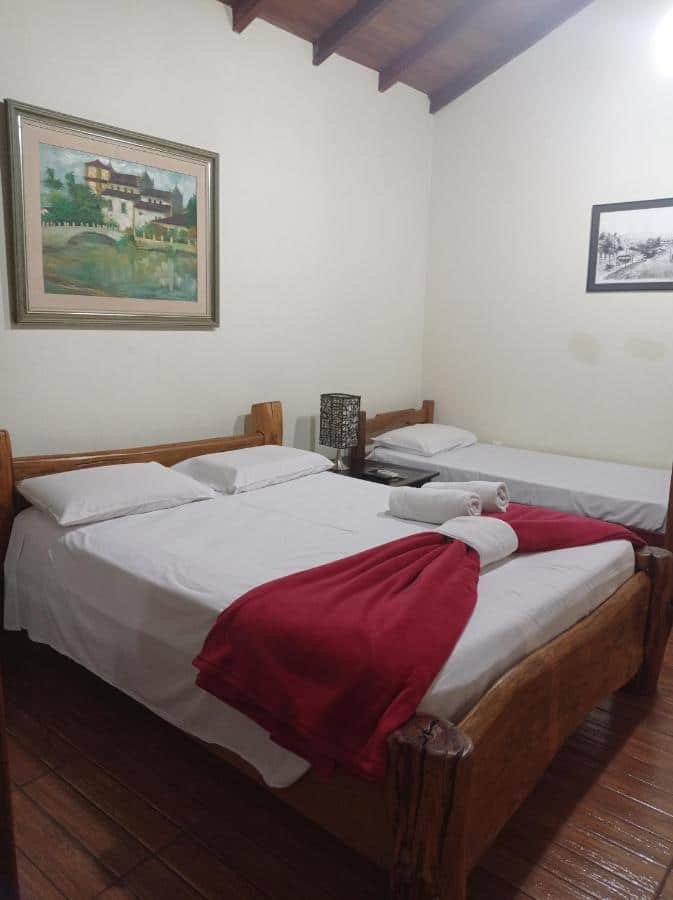 Quarto com uma cama de casal e outra cama de solteiro em Brisa da Serra Hotel Pousada Pirenópolis. Camas cobertas de edredom branco, mesa de cabeceira, abajour, chão de madeira e parede branca com quadro, 