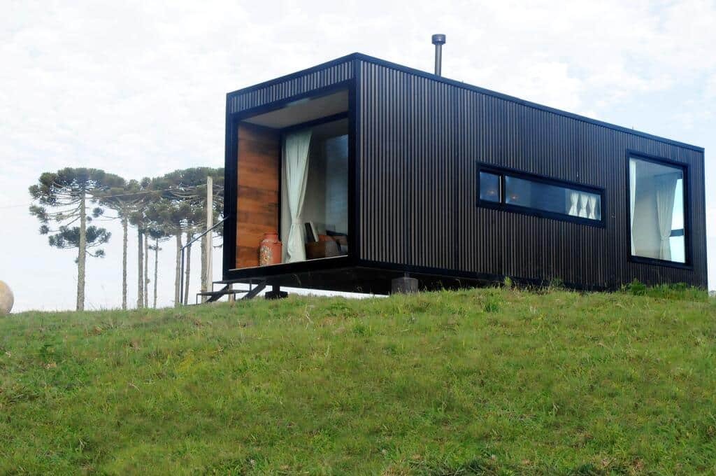 container Brumas Casa de Campo preto com janelas e cortinas brancas, móveis modernos e gramado verde. Ao fundo é possível ver algumas araucárias para trás da casa de temporada