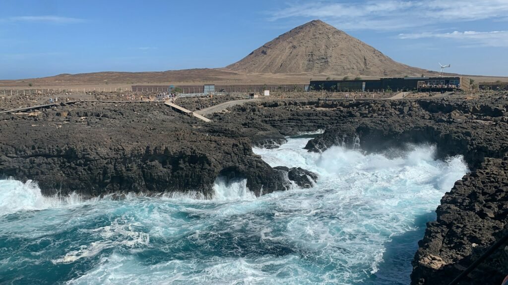 Região de Cabo Verde que se chama Buracona onde mostra as águas fortes batendo nas pedras e ao fundo uma montanha durante o dia. Imagem ilustrando post Hotéis em Cabo Verde.