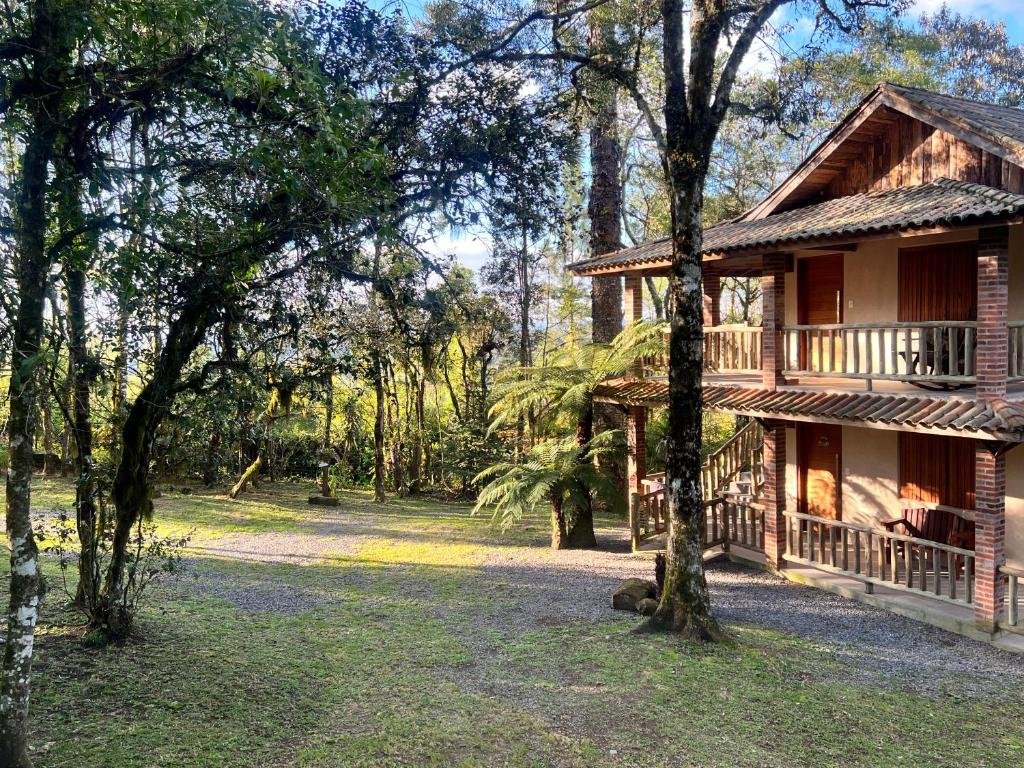 Exterior da Cabana Drechsler Gramado. Há várias árvores e o chalé está na direita da foto, com uma estrutura de dois andares.