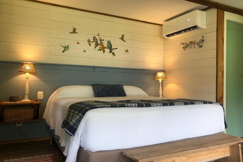 Foto do interior da Cabana Mirim Estalagem Rural representando o post de chalés em Gramado. No quarto vemos uma cama box de casa centralizada na foto. Há um criado-mudo com abajur em seus dois lados e um ar-condicionado está no canto superior direito.