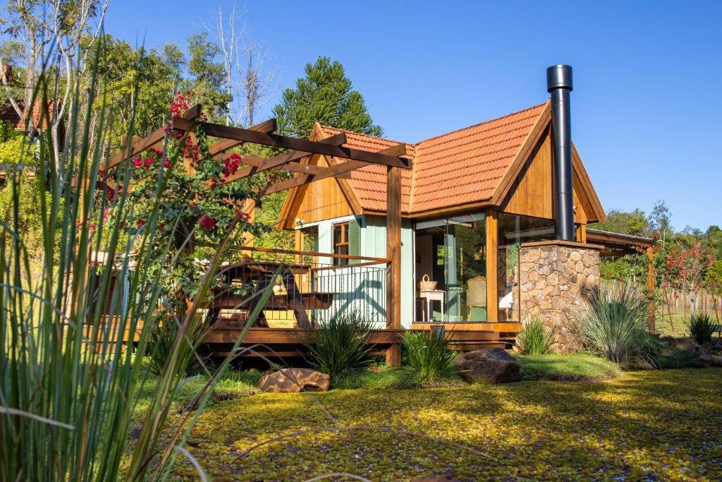 Exterior da Cabana Mirim Estalagem Rural. Há um gramado bem verde e várias flores e árvores. A cabana é de madeira e parte dela está pintada de azul e outra marrom.