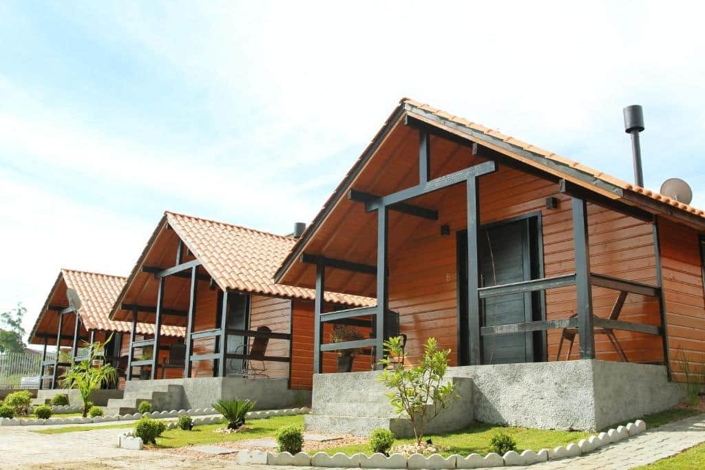 três Cabanas Charme da Serra enfileiradas lado a lado. As três são feitas em madeira com varanda de pedra na frente e detalhes decorativos pintados em preto, como os umbrais das portas e o guarda-corpo. 