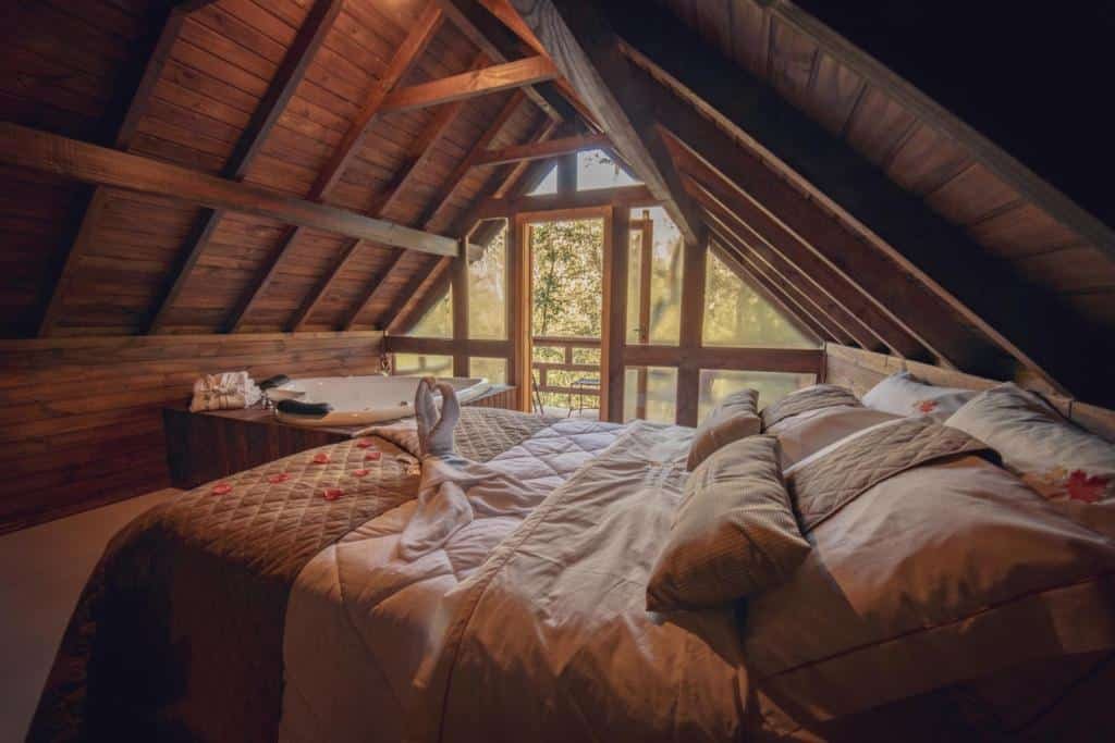 Foto do interior das Cabanas dos Plátanos representando o post de chalés em Gramado. Há uma cama de casal em um chalé que possui um telhado de madeira triangular. No fundo da foto há uma banheira de hidromassagem e uma varanda com vista.