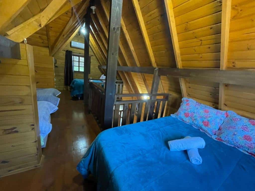 A foto mostra o interior das Cabanas Leão da Tribo de Judá, feitas de madeira. Em primeiro plano, há uma cama com roupa de cama azul e um travesseiro floral. Sobre a cama, há uma toalha branca enrolada. O ambiente possui paredes, piso e teto de madeira, com vigas expostas. À esquerda, é possível ver parcialmente outra cama com roupa de cama semelhante. Ao fundo, há um corrimão de madeira que sugere uma área de loft acima, e através desse corrimão, pode-se ver outra parte da cabana. As janelas permitem a entrada de luz natural, iluminando o ambiente.