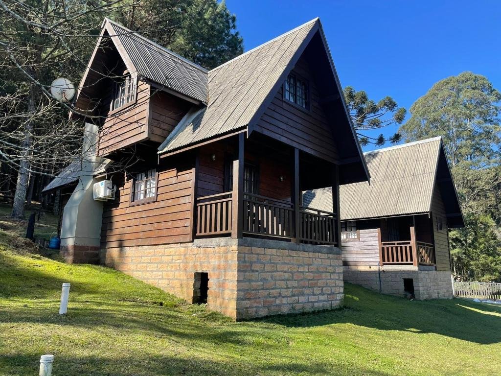 A foto mostra uma cabana de madeira com um telhado inclinado, situada em uma encosta gramada com árvores ao fundo sob um céu azul claro. A cabana tem uma fundação elevada feita de pedra e há várias janelas visíveis nos dois andares. A área ao redor parece bem cuidada, com grama curta e luzes instaladas no chão.