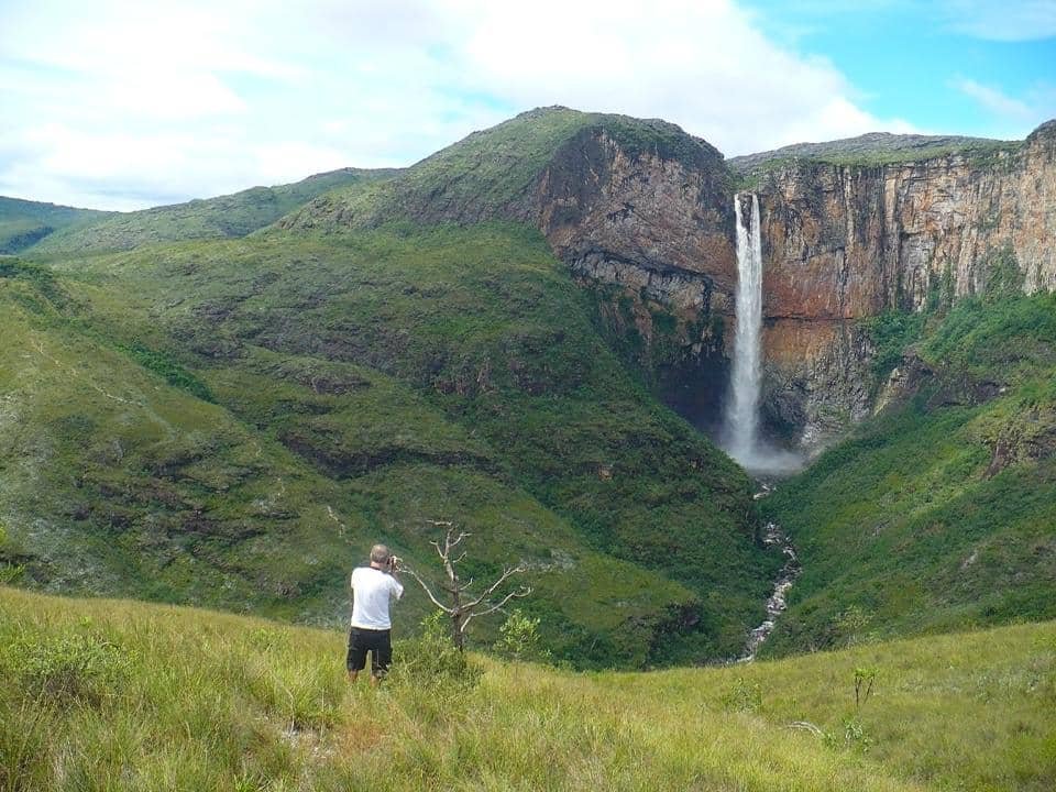 Homem parado em meio ao gramado do parque tirando uma foto da Cachoeira do Tabuleiro ao longe.