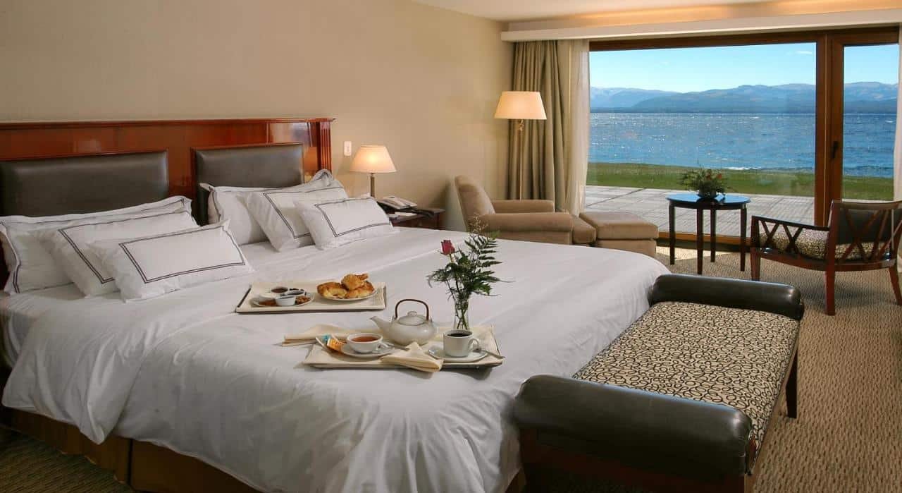 Uma cama de casal do Cacique Inacayal Lake Hotel & Spa com pratos do café da manhã e um sofá. No fundo duas poltronas com mesa de centro com vista do mar.