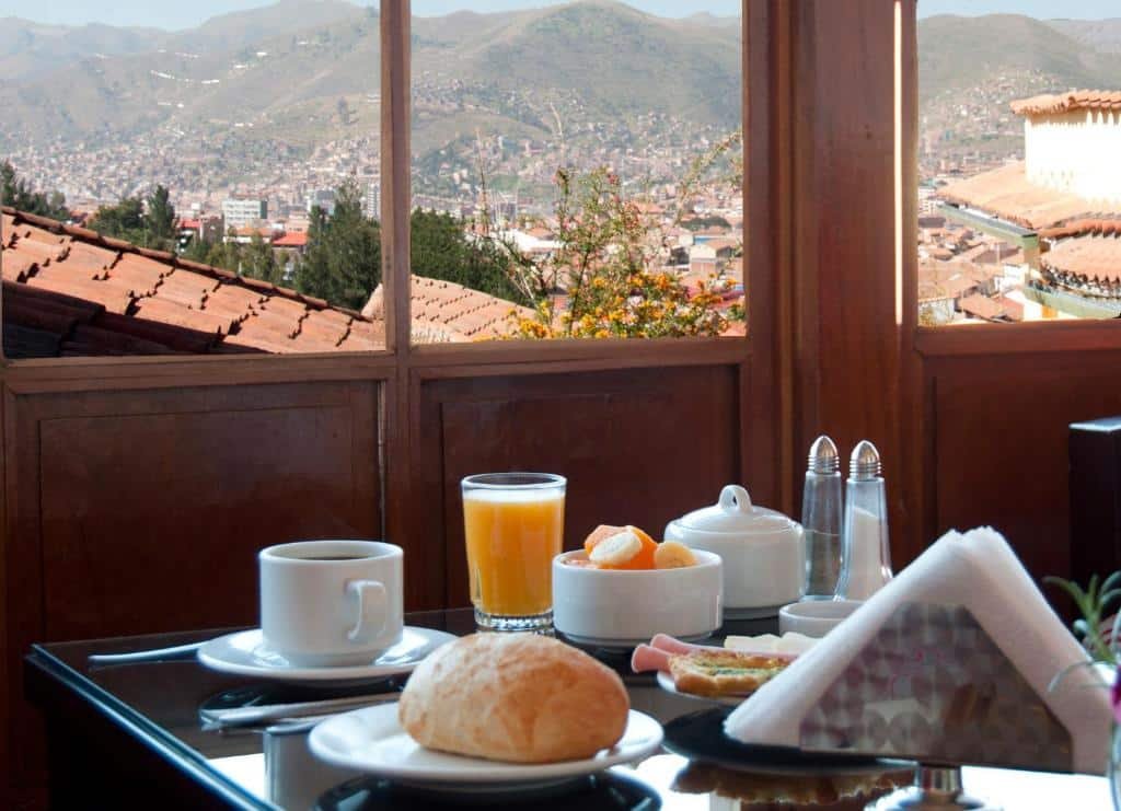 refeitório do Amaru Colonial mostrando uma mesa repleta de pratos e louças brancas, com pães, café, suco de laranja e frutas. Ao fundo há janelas panorâmicas com vista para a cidade.