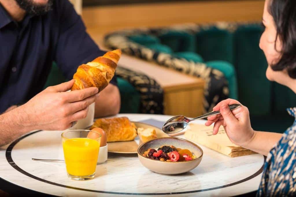 Um homem e uma mulher tomando café da manhã no Mercure Luxemburgo. A Mulher, na direita, segura uma colher enquanto come uma tigela com frutas. O homem, na sua frente, come um croissant, acompanhado de um copo de vidro com suco de laranja. 