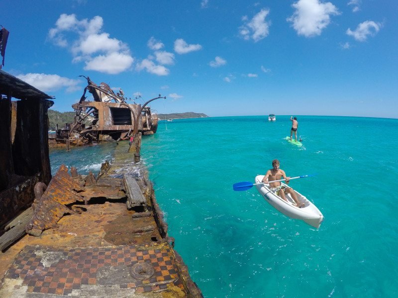Uma pessoa em um caiaque e outra em uma prancha de stand up paddle perto de naufrágios enferrujados em águas azuis cristalinas sob um céu brilhante em Moreton Island
