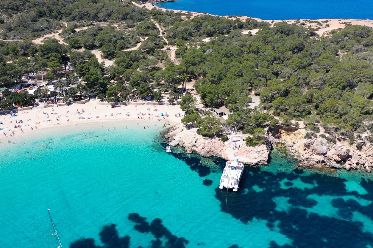 Vista aérea de uma praia de areia com águas azul-turquesa, cercada por árvores e penhascos rochosos, com alguns barcos atracados em um píer. Essa é a Cala Bassa, uma das praias em Ibiza recomendadas.