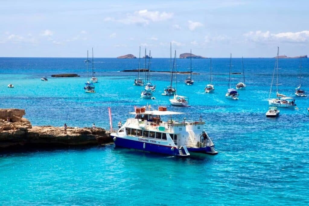 Um barco azul e branco está atracado perto de uma costa rochosa. Vários veleiros estão ancorados no mar azul claro sob um céu ensolarado.