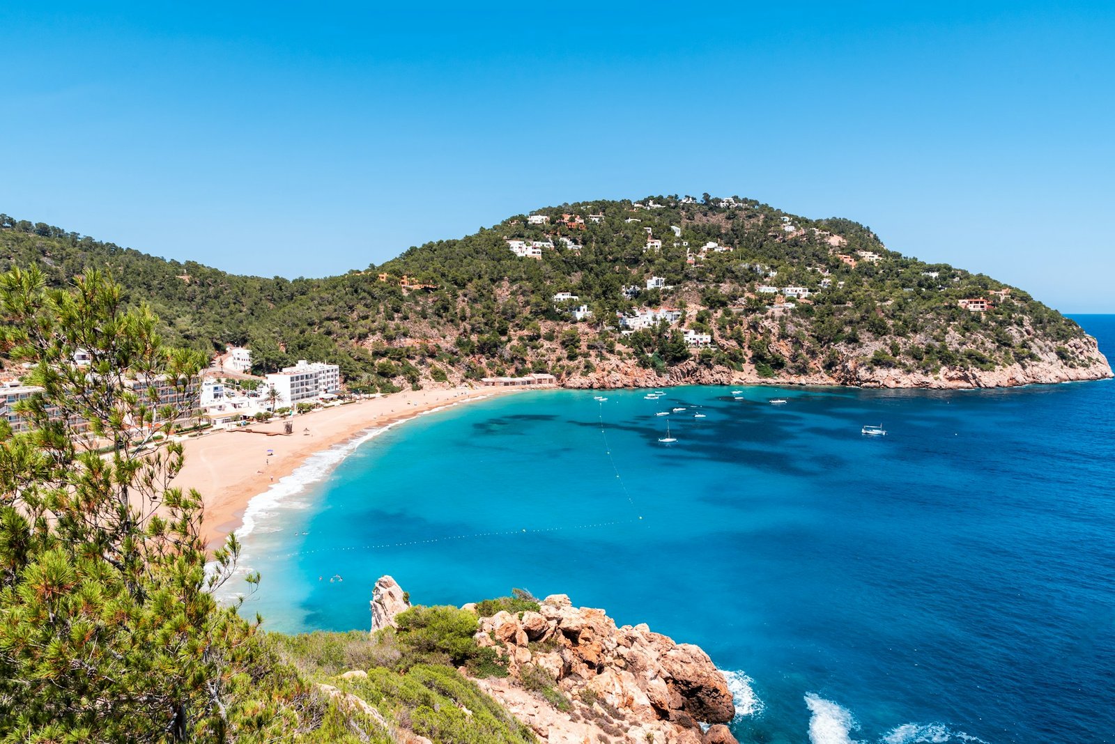 Uma praia com águas azul-turquesa, cercada por colinas verdejantes e vários edifícios, sob um céu azul claro.