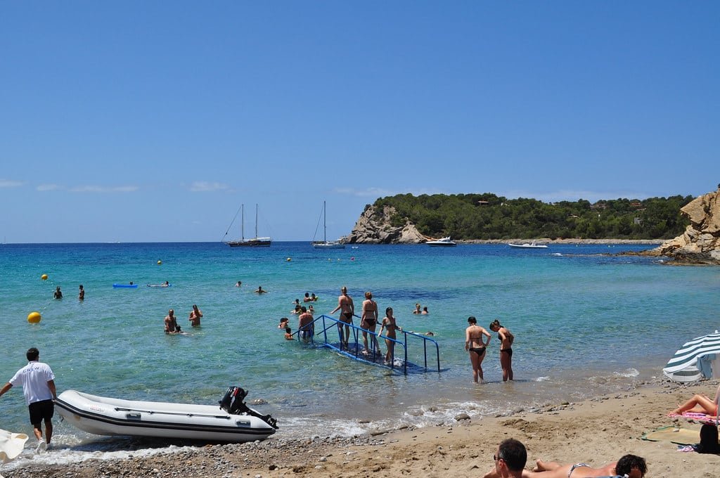 Pessoas aproveitando um dia ensolarado na praia, com nadadores na água e barcos à distância. Uma rampa leva ao mar.