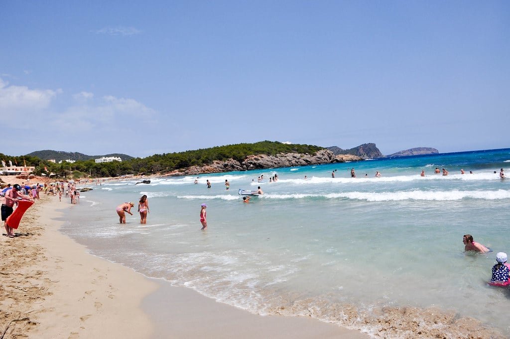 As pessoas aproveitam um dia ensolarado em uma praia arenosa com ondas suaves. Alguns estão nadando enquanto outros relaxam ou caminham pela costa.