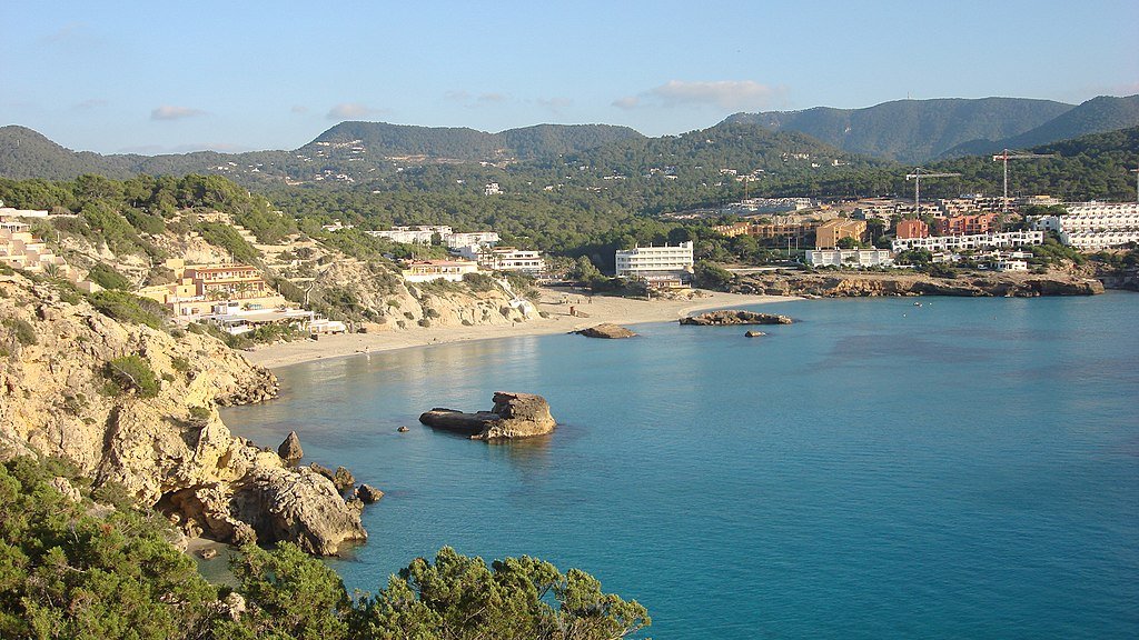 Vista panorâmica de um litoral rochoso com praias de areia, águas azuis cristalinas e colinas verdes sob um céu parcialmente nublado. Essa é a Cala Tarida, uma das praias em Ibiza recomendadas.