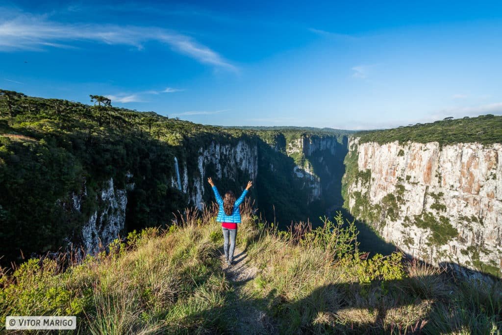 mulher em pé, de costas para a câmera e de frente para o cânion do Itaimbezinho, usando um casaco azul claro, camiseta vermelha e jeans cinza, está com os braços levantados para cima fazendo sinal de paz e amor. À sua frente é possível ver as paredes de pedra dos cânions e um céu azul claro e sem nuvens