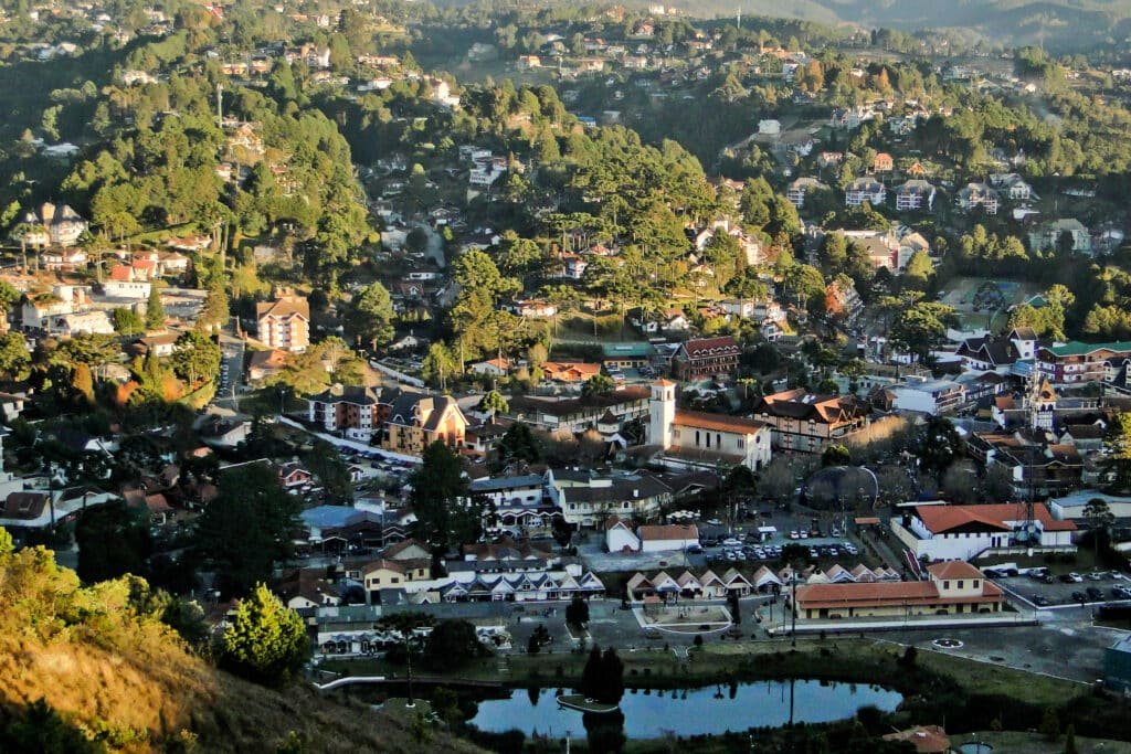 Vista aérea de Campos do Jordão cercada por densas colinas verdes, com casas, uma igreja e um pequeno lago em primeiro plano. Imagem para ilustrar post sobre o que fazer no feriado de Corpus Christi.