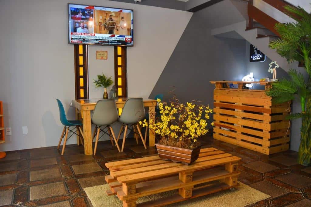 Recepção da CanCoq Guest House, com balcão e mesa de centro rústicos feitos de palets. Há um vaso com flores amarelas em cima da mesinha de centro, e no lado esquerdo há uma mesa retangular encostada na parede com quatro cadeiras. Há também uma TV suspensa na parede passando o jornal da CNN na tela