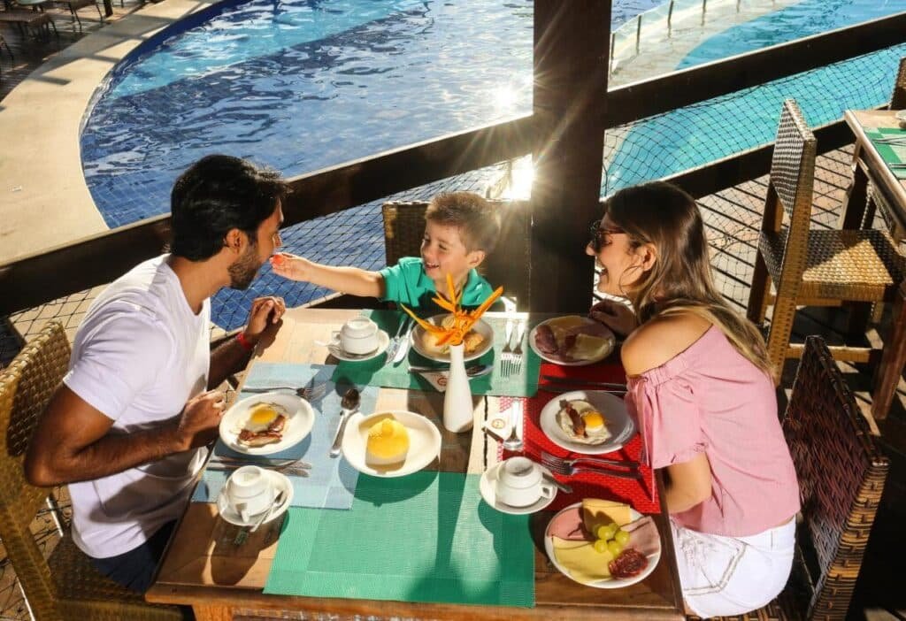 Uma família desfruta do café da manhã na piscina, enquanto uma criança alimenta um homem. A luz do sol brilha na mesa posta com café e itens de café da manhã no Cana Brava All Inclusive Resort.