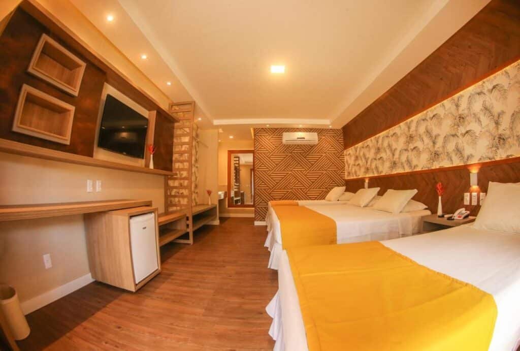 Quarto do Cana Brava All Inclusive Resort com duas camas de casal, cabeceiras amarelas, decoração em madeira, TV na parede, escrivaninha, frigobar e arte decorativa na parede.