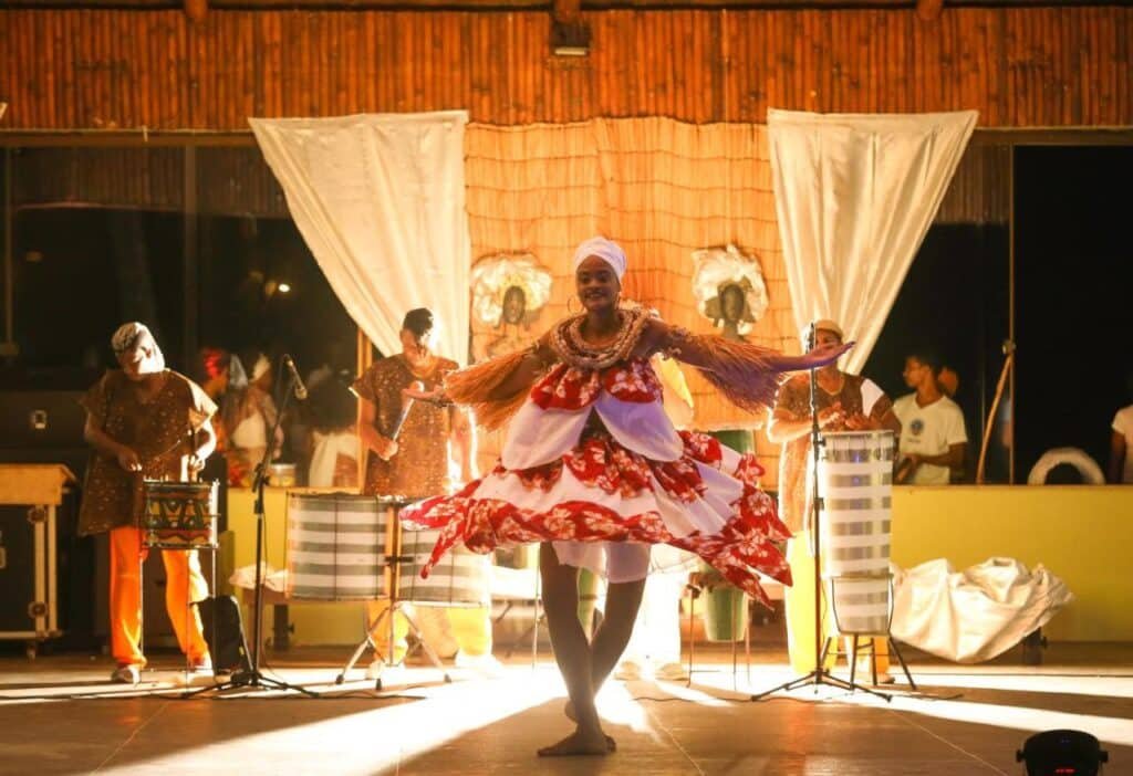 Uma dançarina com um vestido colorido se apresenta no palco com uma banda ao vivo ao fundo, iluminada por uma iluminação quente no Cana Brava All Inclusive Resort.