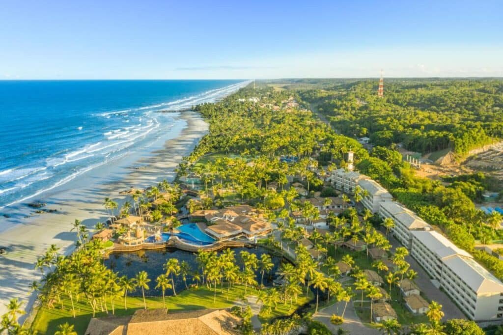 Vista aérea do Cana Brava All Inclusive Resort costeiro com edifícios, piscinas e palmeiras perto de uma praia com ondas sob um céu azul claro.