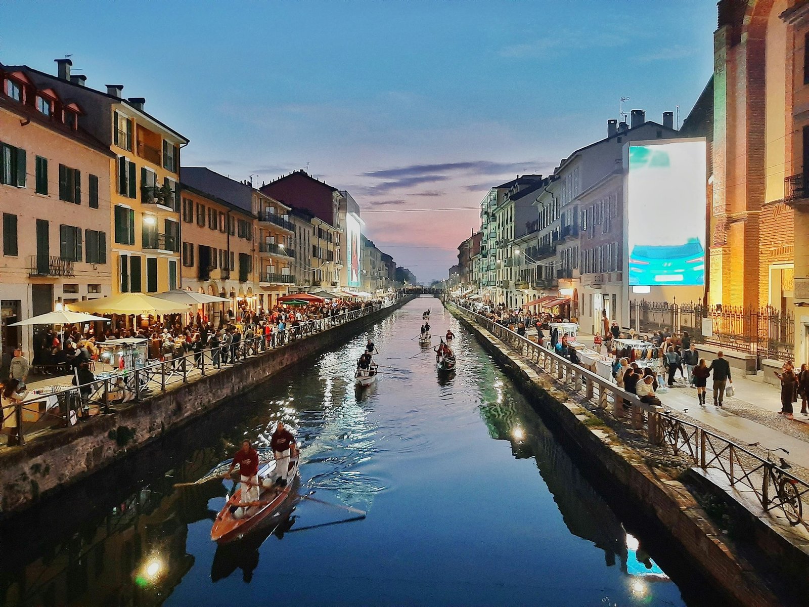 Canal em Navigli alinhado com prédios e cafés sob um céu crepuscular. Barcos com pessoas estão na água, e há uma grande tela digital. Foto para o post de o que faer em Milão.