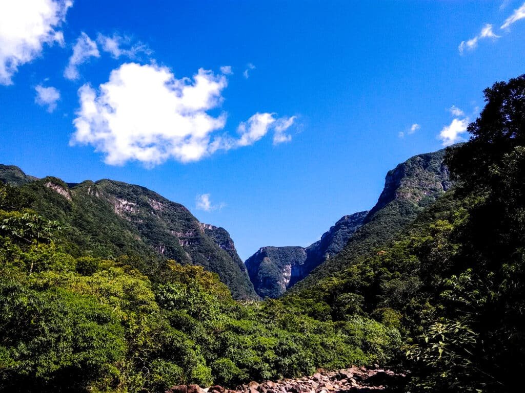 Vale verdejante com montanhas rochosas sob um céu azul brilhante e nuvens dispersas.