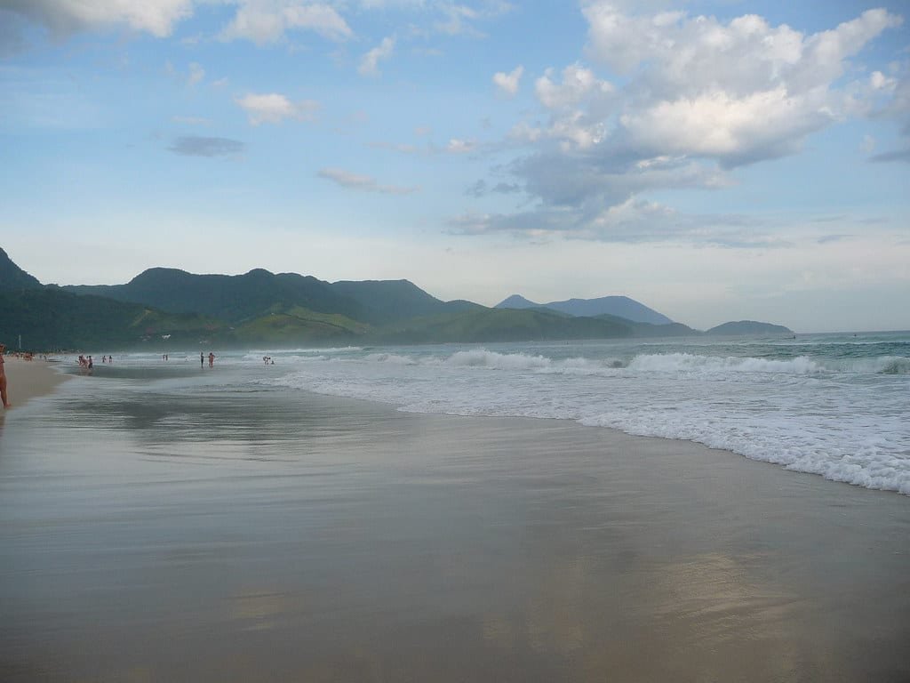 Uma praia calma com ondas suaves batendo na praia, montanhas distantes ao fundo e algumas pessoas caminhando na areia. Essa é a praia de Maresias em São Sebastião, no Litoral Norte de São Paulo.
