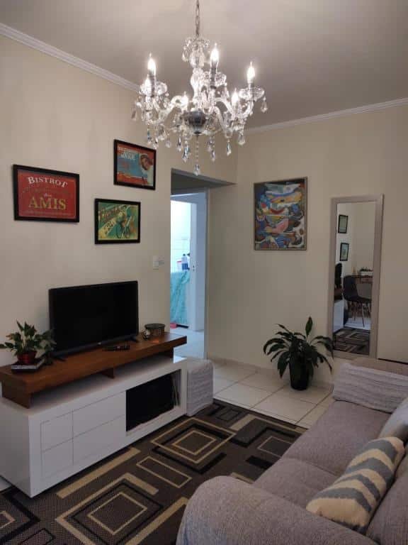 Sala de estar em Casa agradável com churrasqueira no centro de Serra Negra. O teto possui um lustre prata. Na direita está o sofá, e de frente para ele há um rack com TV, e atrás há uma parede com quadros. Há duas portas que levam a diferentes cômodos no fundo da imagem.