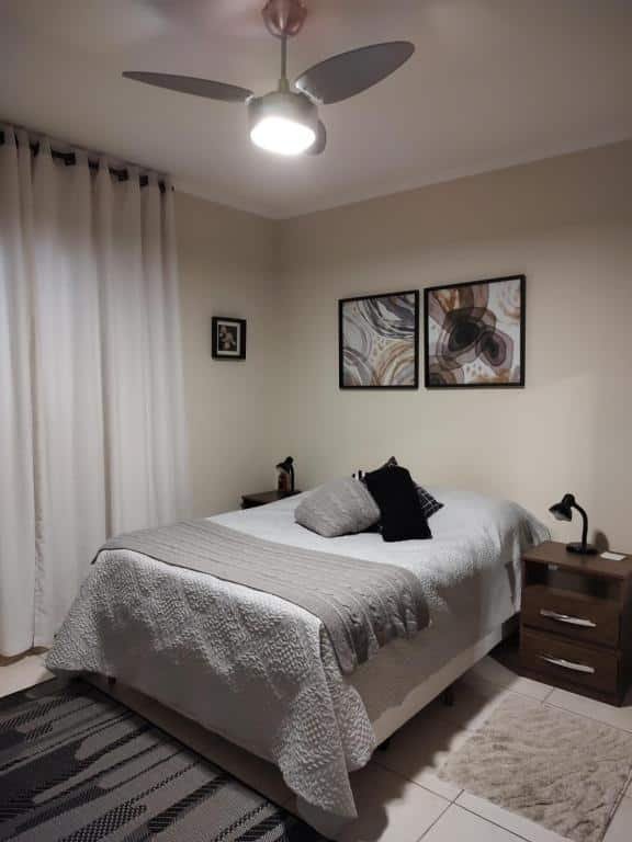 Foto do quarto em Casa agradável com churrasqueira no centro de Serra Negra. Há uma cama de casal ao centro. De seus dois lados há uma mesa de cabeceira de madeira com abajur em cima. Na parede atrás da cama há dois quadros decorativo. Na esquerda há uma janela com cortinas brancas.
Ilustra o post sobre airbnb em Serra Negra.
