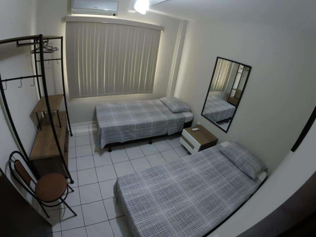 Quarto da Casa Área Central. Do lado direito duas camas de solteiro, entre elas uma estante e um espelho. Do lado esquerdo um armário e uma cadeira. No fundo a janela do quarto com cortina fechada.