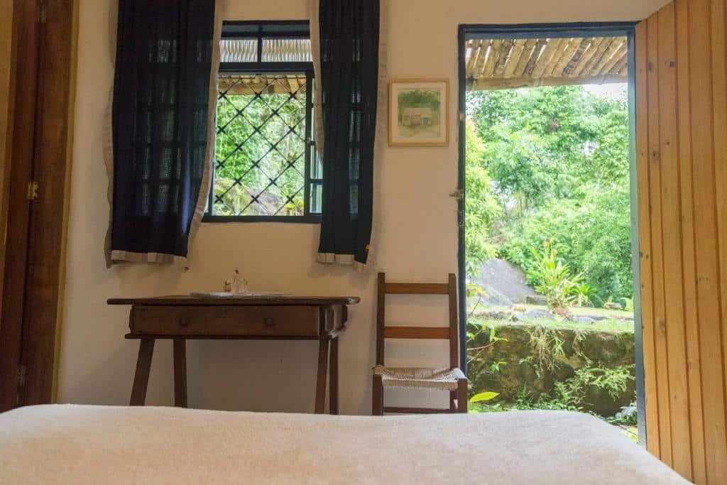 Quarto da Casa das Pedras. Uma cama de casal no meio, de frente uma mesa, uma cadeira, a janela do quarto e a porta com vista para a mata. Foto para ilustrar post sobre airbnb em Ubatumirim.