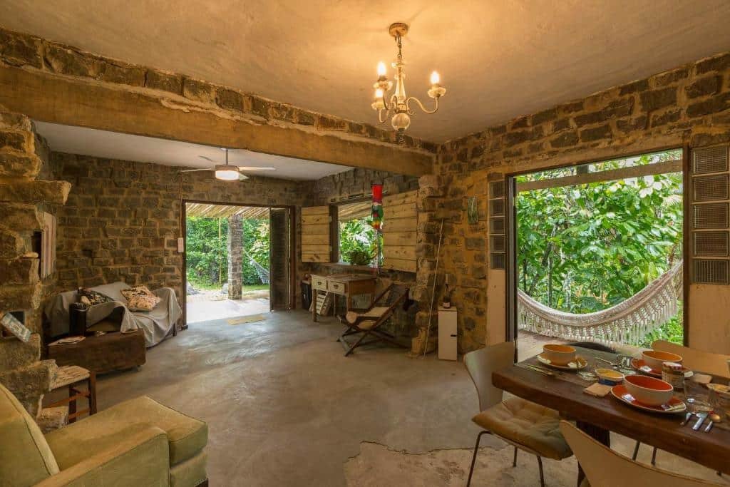 Sala de estar da Casa das Pedras. Do lado direito uma mesa posta, do lado esquerdo uma poltrona, no meio cadeiras e sofás, e ao redor portas grandes com vista para a mata e uma rede.