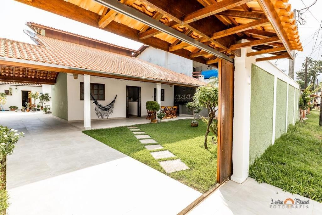 Entrada da Casa Flor. Um portão grande está aberto e mostra a entrada, onde há um espaço para estacionar. Logo ao lado dele está a casa com uma porta, duas janelas e uma rede. Uma parte do quintal é toda de grama.