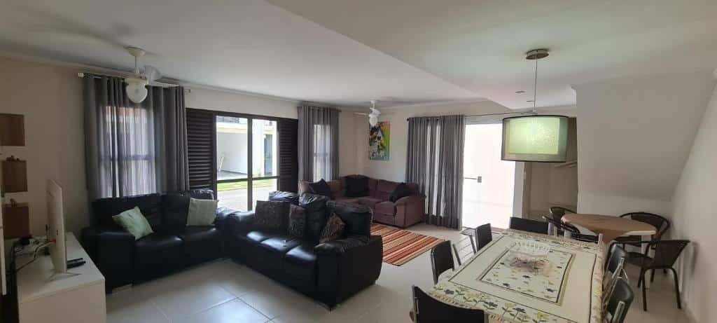 Sala de estar da Casa Melo 4. Duas mesas do lado direito, do lado esquerdo uma raque com televisão e três sofás. Do lado esquerdo e no fundo porta da casa.