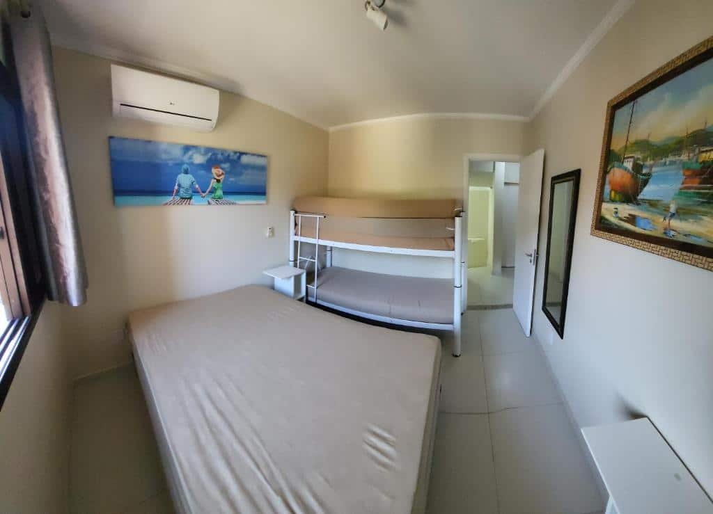 Quarto da Casa Melo 4. Uma cama de casal do lado esquerdo, no fundo uma cama de beliche. Em cima das camas um quadro e um ar-condicionado. No lado direito um quadro, um espelho e a porta do quarto aberta. Foto para ilustrar post sobre airbnb em Perequê Mirim.