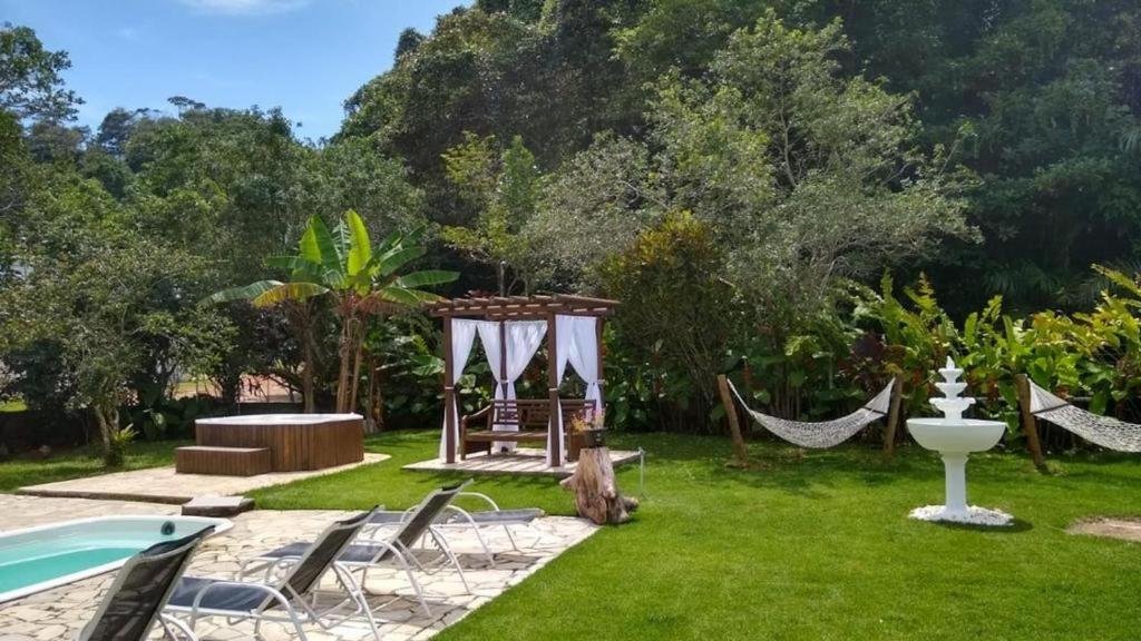 Jardim da Casa Pé na Areia em Ubatuba – Alto Padrão a 30m da Praia. Uma piscina está no canto com algumas espreguiçadeiras ao lado. Ao fundo há um banco, um ofurô, algumas redes e uma pequena fonte. O ambiente é cercado de árvores.