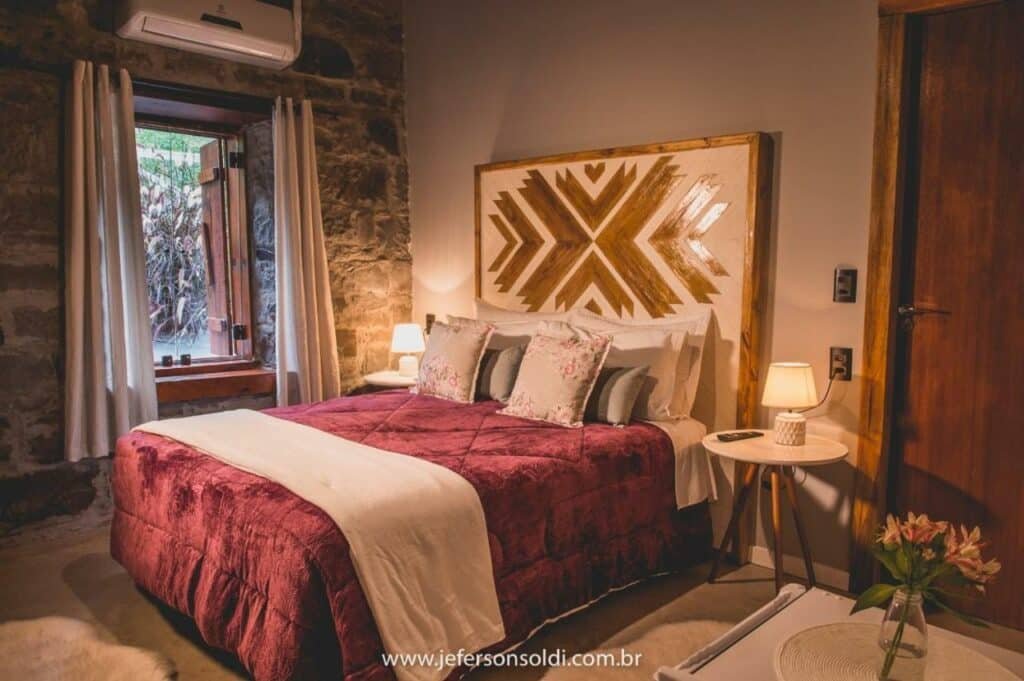 Cama de casal da Casa Vêneto com colcha de aparência aveludada na cor vermelha, cabeceira alta de madeira e almofadas cor nude, uma janela aberta ao fundo, com cortina, e mesinha de cabeceira com abajures em casa lado da cama