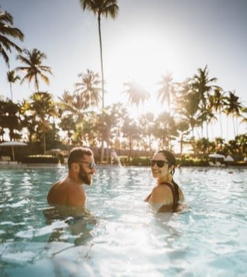 Um homem e uma mulher usando óculos escuros estão na piscina do TRS Cap Cana Waterfront & Marina Hotel, sorrindo um para o outro, com palmeiras e um sol brilhante ao fundo para representar um dos melhores lugares para lua de mel