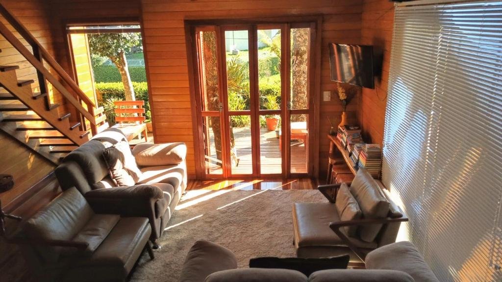 Imagem da sala na Casas Ana. As paredes são de madeira, Há dois sofás, um à esquerda e outro no canto inferior direito. Atrás do da esquerda, há uma escada. A tv está na parede, de frente para os sofás, na diagonal. Há uma porta de vidro no fundo que leva para a varanda.